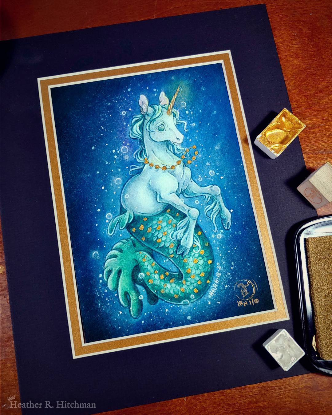 Photograph of a matted print of a full body watercolor, gouache, and colored pencil painting of a mermaid unicorn under the sea. Also known as a mermacorn, the creature has a white unicorn torso and a green mermaid tail. The mermacorn appears to be a young colt, with large light green eyes, a short glowing golden horn, and long spindly front legs. The unicorn wears a necklace of golden pearls and floats effortlessly in the dark waters with a smile on its face. They are facing ¾ to the right, and appear to be glowing so that the dark waters immediately around them are illuminated to a lighter blue color. The mermacorn has golden scales scattered among the length of its green tail. The background is a gradient of blue water, with white bubbles surrounding the floating mermacorn from head to tail. The mat-board is black on the top layer and gold on the second layer. Small pans of gold and silver watercolor pigment and a golden ink pad surround the print.