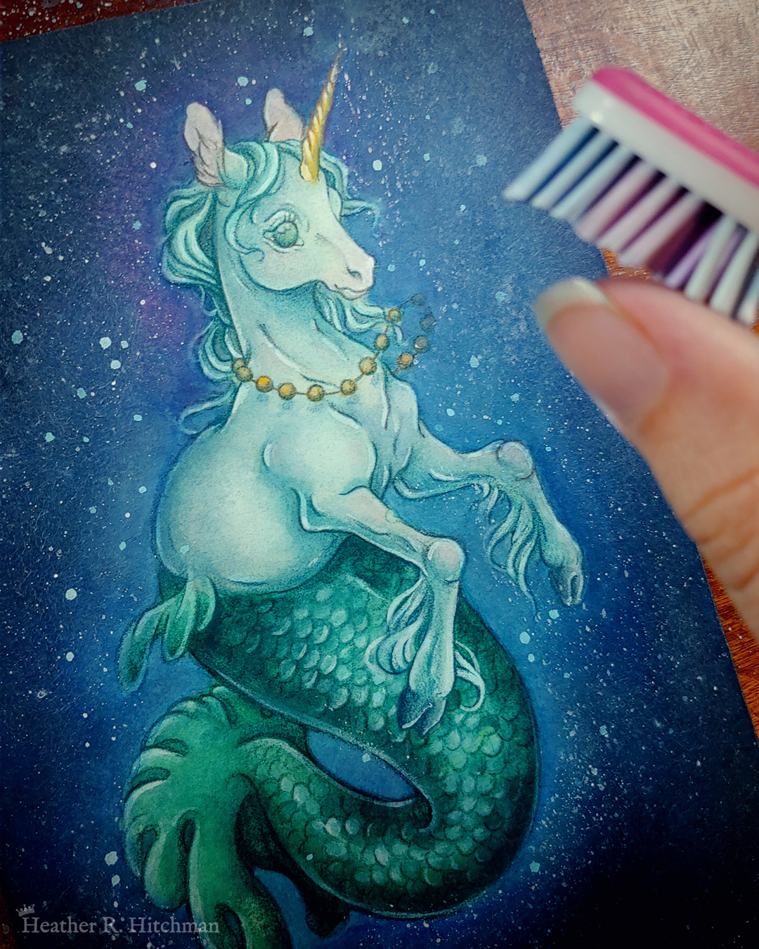 Process photograph of a full body watercolor, gouache, and colored pencil painting of a mermaid unicorn under the sea. Also known as a mermacorn, the creature has a white unicorn torso and a green mermaid tail. The mermacorn appears to be a young colt, with large light green eyes, a short glowing golden horn, and long spindly front legs. The unicorn wears a necklace of golden pearls and floats effortlessly in the dark waters with a smile on its face. They are facing ¾ to the right, and appear to be glowing so that the dark waters immediately around them are illuminated to a lighter blue color. The mermacorn has golden scales scattered among the length of its green tail. The background is a gradient of blue water, with white bubbles surrounding the floating mermacorn from head to tail. The artist’s hand is in the top right corner using a pink toothbrush to flick spatters of white paint onto the painting, creating misty texture in the water.