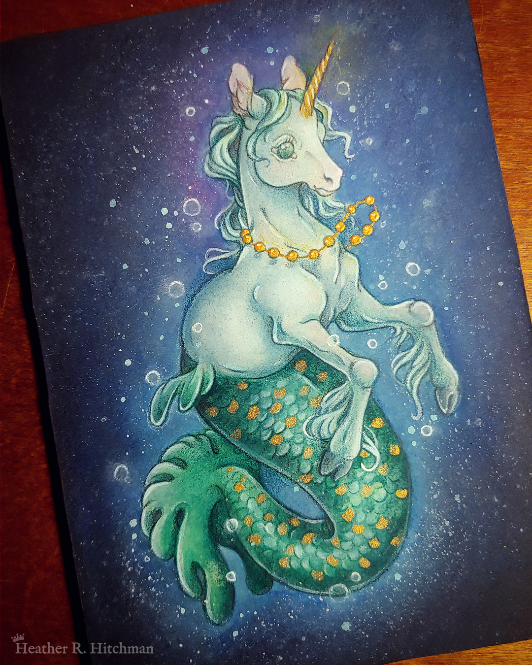 Photograph of a full body watercolor, gouache, and colored pencil painting of a mermaid unicorn under the sea. Also known as a mermacorn, the creature has a white unicorn torso and a green mermaid tail. The mermacorn appears to be a young colt, with large light green eyes, a short glowing golden horn, and long spindly front legs. The unicorn wears a necklace of golden pearls and floats effortlessly in the dark waters with a smile on its face. They are facing ¾ to the right, and appear to be glowing so that the dark waters immediately around them are illuminated to a lighter blue color. The mermacorn has golden scales scattered among the length of its green tail. The background is a gradient of blue water, with white bubbles surrounding the floating mermacorn from head to tail. The painting is resting in a slanted position on a wooden tabletop.