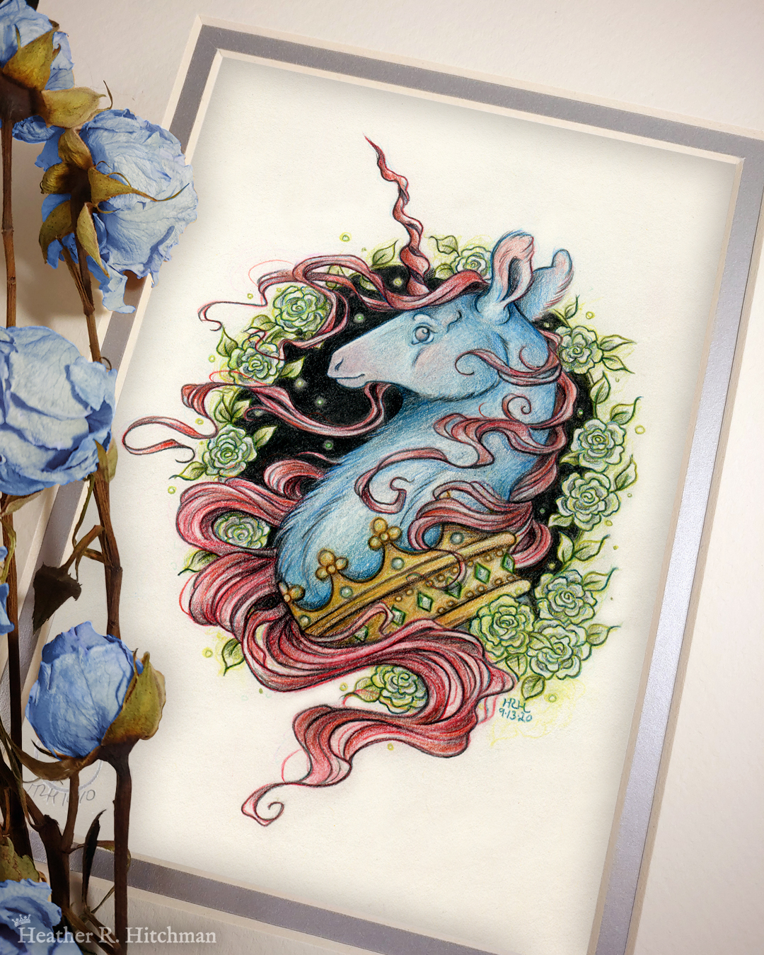 Photograph of a matted print of a graphite and colored pencil portrait drawing of a blue and red dark unicorn bust framed in a circular wreath of soft green gardenia flowers. The background inside of the wreath behind the unicorn is sepia with tiny orbs of green light floating around. Outside of the wreath the portrait has a sepia background color. The unicorn lifts his head proudly in profile facing to the left. He has a flowing red mane and chin beard, blue fur, and a long red corkscrew horn in the center of his forehead. The dark unicorn wears a large crown of emerald and ruby jewels around his neck. The mat-board is white on the top layer and silver on the second layer. Blue roses rest on top of the print to the left. A small silver stamp with the Terratoff icon is at the bottom left corner of the print, under the roses.