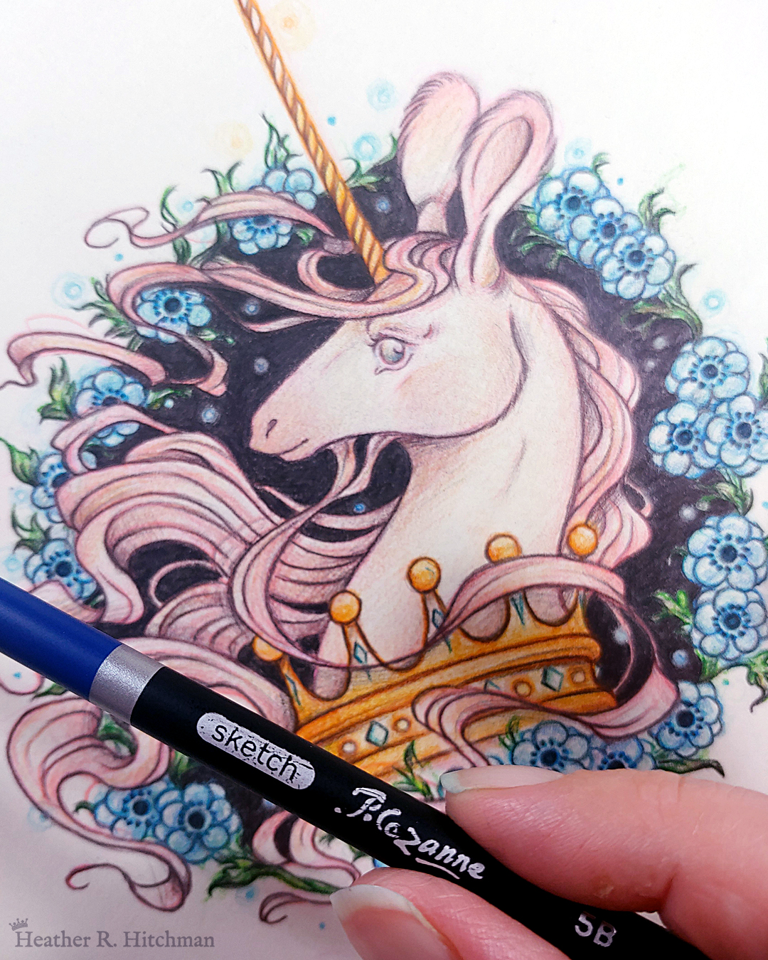 Process photograph of a graphite and colored pencil portrait drawing of a light pink colored swift unicorn bust framed in a circular wreath of blue anemone flowers. The background inside of the wreath behind the unicorn is black with tiny orbs of blue light floating around. Outside of the wreath the portrait has a sepia background color. The unicorn lifts her head in profile facing to the left with a gentle expression on her face. She has a flowing pink mane and chin beard, light pink fur, and an extremely long golden horn in the center of her forehead. The unicorn wears a large crown of blue and pink jewels around her neck. The artist’s hand is in the lower right corner holding up a graphite pencil over the artwork.