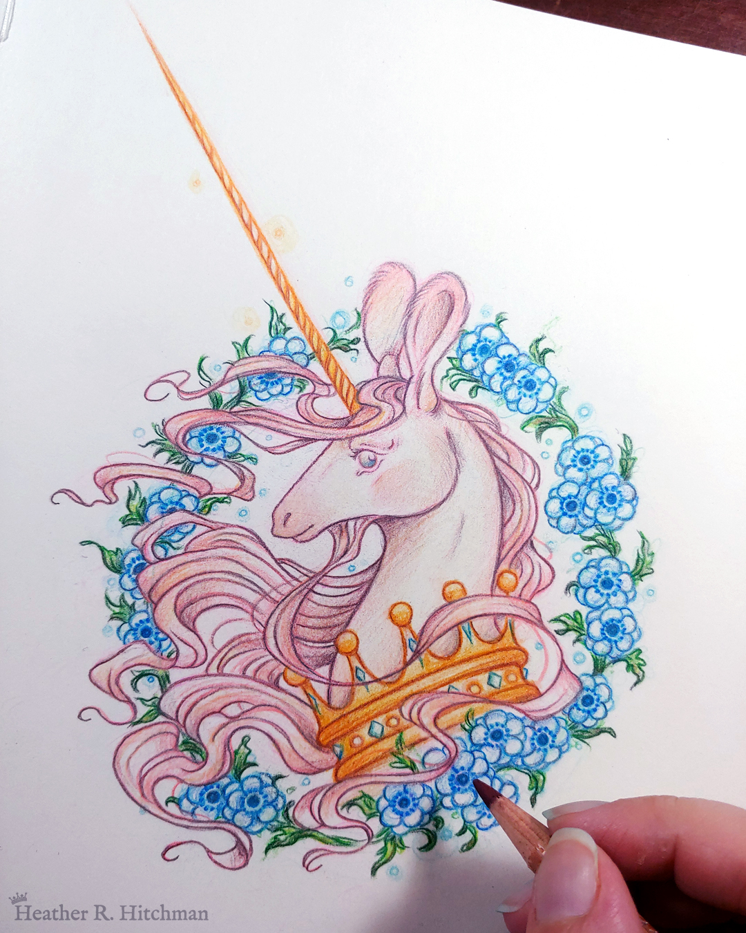 Process photograph of a graphite and colored pencil portrait drawing of a light pink colored swift unicorn bust framed in a circular wreath of blue anemone flowers. The background inside of the wreath behind the unicorn is white with tiny orbs of blue light floating around. Outside of the wreath the portrait has a white background color. The unicorn lifts her head in profile facing to the left with a gentle expression on her face. She has a flowing pink mane and chin beard, light pink fur, and an extremely long golden horn in the center of her forehead. The unicorn wears a large crown of blue and pink jewels around her neck. The artist’s fingertips are in the lower right corner holding the tip of a graphite pencil over the artwork.