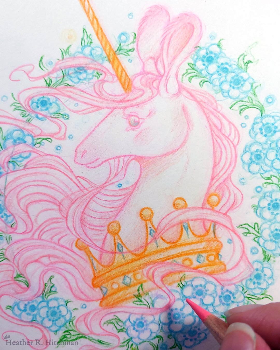 Process photograph of a graphite and colored pencil portrait drawing of a light pink colored swift unicorn bust framed in a circular wreath of blue anemone flowers. The background inside of the wreath behind the unicorn is white with tiny orbs of blue light floating around. Outside of the wreath the portrait has a white background color. The unicorn lifts her head in profile facing to the left with a gentle expression on her face. She has a flowing pink mane and chin beard, light pink fur, and an extremely long golden horn in the center of her forehead. The unicorn wears a large crown of blue and pink jewels around her neck. The artist’s fingertips are in the lower right corner holding the tip of a pink pencil over the artwork.