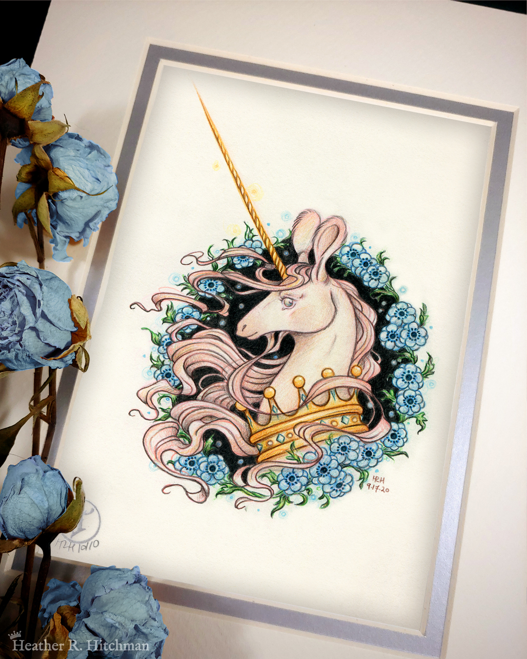 Photograph of a matted print of a graphite and colored pencil portrait drawing of a light pink colored swift unicorn bust framed in a circular wreath of blue anemone flowers. The background inside of the wreath behind the unicorn is black with tiny orbs of blue light floating around. Outside of the wreath the portrait has a sepia background color. The unicorn lifts her head in profile facing to the left with a gentle expression on her face. She has a flowing pink mane and chin beard, light pink fur, and an extremely long golden horn in the center of her forehead. The unicorn wears a large crown of blue and pink jewels around her neck. The mat-board is white on the top layer and silver on the second layer. Blue roses rest on top of the print to the left. A small silver stamp with the Terratoff icon is at the bottom left corner of the print, under the roses.