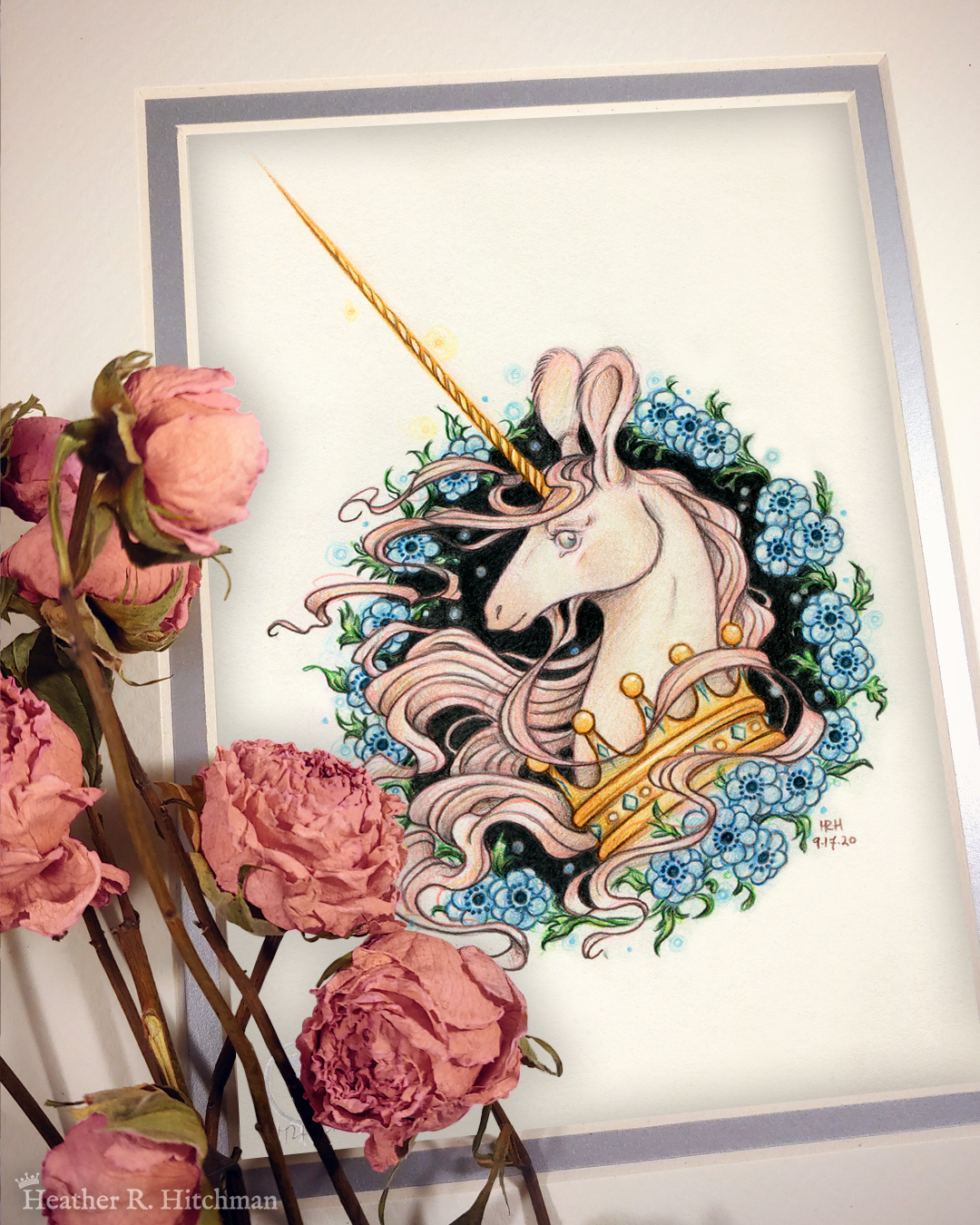Photograph of a matted print of a graphite and colored pencil portrait drawing of a light pink colored swift unicorn bust framed in a circular wreath of blue anemone flowers. The background inside of the wreath behind the unicorn is black with tiny orbs of blue light floating around. Outside of the wreath the portrait has a sepia background color. The unicorn lifts her head in profile facing to the left with a gentle expression on her face. She has a flowing pink mane and chin beard, light pink fur, and an extremely long golden horn in the center of her forehead. The unicorn wears a large crown of blue and pink jewels around her neck. The mat-board is white on the top layer and silver on the second layer. Pink roses rest on top of the print to the left. A small silver stamp with the Terratoff icon is at the bottom left corner of the print, under the roses.