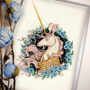 Photograph of a matted print of a graphite and colored pencil portrait drawing of a light pink colored swift unicorn bust framed in a circular wreath of blue anemone flowers. The background inside of the wreath behind the unicorn is black with tiny orbs of blue light floating around. Outside of the wreath the portrait has a sepia background color. The unicorn lifts her head in profile facing to the left with a gentle expression on her face. She has a flowing pink mane and chin beard, light pink fur, and an extremely long golden horn in the center of her forehead. The unicorn wears a large crown of blue and pink jewels around her neck. The mat-board is white on the top layer and silver on the second layer. Blue roses rest on top of the print to the left. A small silver stamp with the Terratoff icon is at the bottom left corner of the print, under the roses.
