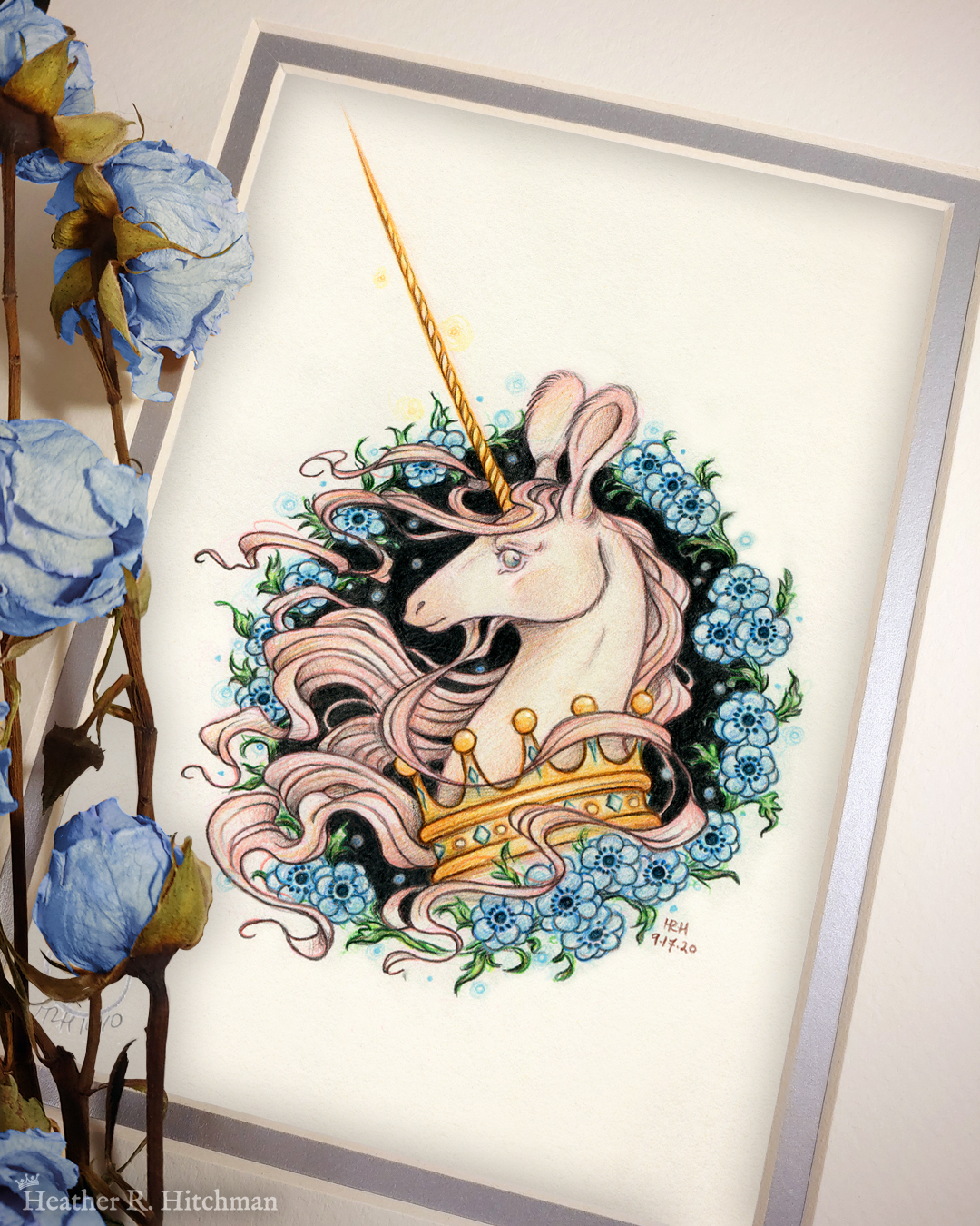 Photograph of a matted print of a graphite and colored pencil portrait drawing of a light pink colored swift unicorn bust framed in a circular wreath of blue anemone flowers. The background inside of the wreath behind the unicorn is black with tiny orbs of blue light floating around. Outside of the wreath the portrait has a sepia background color. The unicorn lifts her head in profile facing to the left with a gentle expression on her face. She has a flowing pink mane and chin beard, light pink fur, and an extremely long golden horn in the center of her forehead. The unicorn wears a large crown of blue and pink jewels around her neck. The mat-board is white on the top layer and silver on the second layer. Blue roses rest on top of the print to the left. A small silver stamp with the Terratoff icon is at the bottom left corner of the print, under the roses.