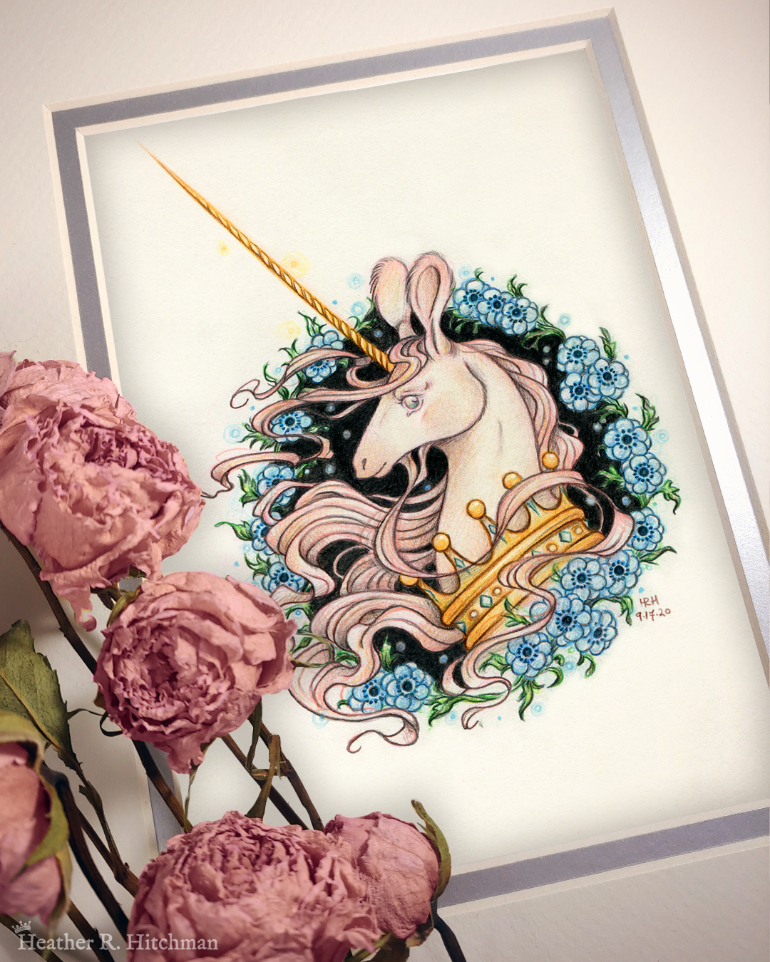 Photograph of a matted print of a graphite and colored pencil portrait drawing of a light pink colored swift unicorn bust framed in a circular wreath of blue anemone flowers. The background inside of the wreath behind the unicorn is black with tiny orbs of blue light floating around. Outside of the wreath the portrait has a sepia background color. The unicorn lifts her head in profile facing to the left with a gentle expression on her face. She has a flowing pink mane and chin beard, light pink fur, and an extremely long golden horn in the center of her forehead. The unicorn wears a large crown of blue and pink jewels around her neck. The mat-board is white on the top layer and silver on the second layer. Pink roses rest on top of the print to the left. A small silver stamp with the Terratoff icon is at the bottom left corner of the print, under the roses.