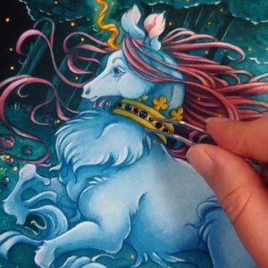 Close up photograph of a watercolor, gouache, and colored pencil painting print of a unicorn running through a moonlit forest. The unicorn is visible from the knees up, and has blue fur that flows in elegant tufts from their chest and hooves. He has a mane of raspberry red that swirls around in the wind behind the unicorn’s head. The unicorn has a twisting golden horn, and wears a golden crown encrusted with red and blue jewels around their neck. The fingertips of the artist are hovering to the right over the crown holding tiny tweezers that she uses to adhere flat back crystals onto the painted crown. Running in profile to the left with their front hooves close to their chest, the unicorn has a peaceful expression on its face. The background is a cool greenish blue night forest scene. In the field red poppies bloom. Trees with dark branches are in the far background, with an opening behind the unicorn that reveals a large full moon glowing white. The scene is sprinkled with tiny golden orbs of light.