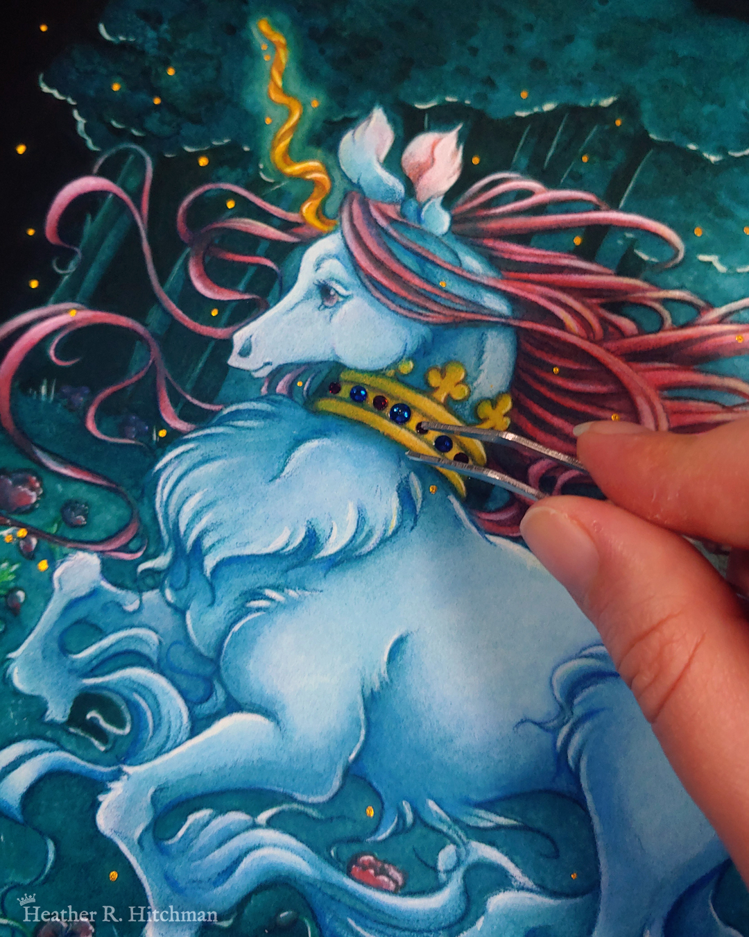 Close up photograph of a watercolor, gouache, and colored pencil painting print of a unicorn running through a moonlit forest. The unicorn is visible from the knees up, and has blue fur that flows in elegant tufts from their chest and hooves. He has a mane of raspberry red that swirls around in the wind behind the unicorn’s head. The unicorn has a twisting golden horn, and wears a golden crown encrusted with red and blue jewels around their neck. The fingertips of the artist are hovering to the right over the crown holding tiny tweezers that she uses to adhere flat back crystals onto the painted crown. Running in profile to the left with their front hooves close to their chest, the unicorn has a peaceful expression on its face. The background is a cool greenish blue night forest scene. In the field red poppies bloom. Trees with dark branches are in the far background, with an opening behind the unicorn that reveals a large full moon glowing white. The scene is sprinkled with tiny golden orbs of light.