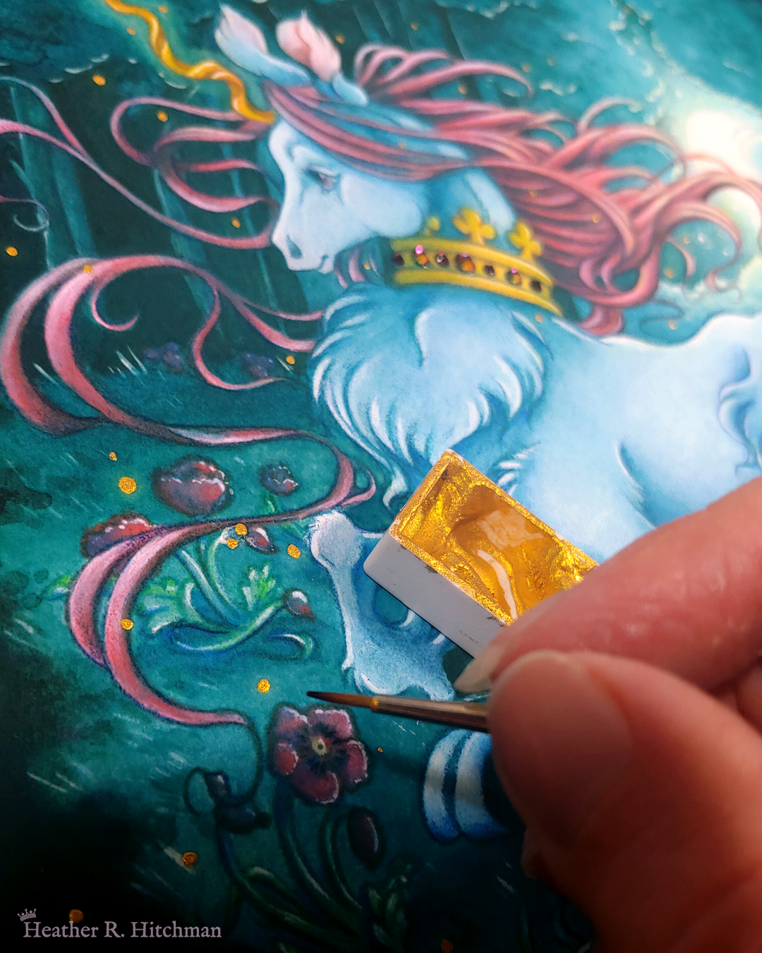 Close up photograph of a watercolor, gouache, and colored pencil painting print of a unicorn running through a moonlit forest. The unicorn is visible from the knees up, and has blue fur that flows in elegant tufts from their chest and hooves. He has a mane of raspberry red that swirls around in the wind behind the unicorn’s head. The unicorn has a twisting golden horn, and wears a golden crown encrusted with red and blue jewels around their neck. Running in profile to the left with their front hooves close to their chest, the unicorn has a peaceful expression on its face. The background is a cool greenish blue night forest scene. In the field red poppies bloom. Trees with dark branches are in the far background, with an opening behind the unicorn that reveals a large full moon glowing white. The scene is sprinkled with tiny golden orbs of light. The fingertips of the artist are hovering to the bottom right corner of the photograph holding a tiny paintbrush, painting on embellished orbs of gold onto the print. A small rectangular pan of golden paint in on top of the painting behind her fingers.