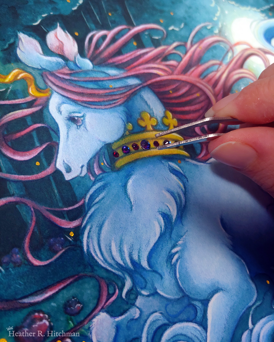 Close up photograph of a watercolor, gouache, and colored pencil painting print of a unicorn running through a moonlit forest. The unicorn is visible from the chest up, and has blue fur that flows in elegant tufts from their chest. He has a mane of raspberry red that swirls around in the wind behind the unicorn’s head. The unicorn has a twisting golden horn, and wears a golden crown encrusted with red and blue jewels around their neck. The fingertips of the artist are hovering to the right over the crown holding tiny tweezers that she uses to adhere flat back crystals onto the painted crown. Running in profile to the left with their front hooves close to their chest, the unicorn has a peaceful expression on its face. The background is a cool greenish blue night forest scene. In the field red poppies bloom. Trees with dark branches are in the far background, with an opening behind the unicorn that reveals a large full moon glowing white. The scene is sprinkled with tiny golden orbs of light.