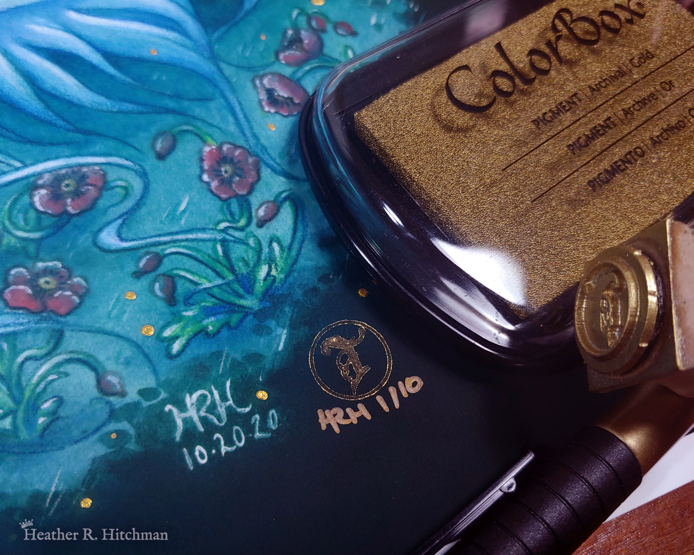 Close up photograph of the bottom right corner of a unicorn painting print by Heather R. Hitchman called “Moonlit Run”. The corner shows the red poppies growing in a field of bluish green grass in a night forest scene. Next to the original artist’s signature there is a golden stamp of the Terratoff icon that has been signed and numbered in gold. Resting on the right side print is the golden ink pad used to make the stamp, a golden pigment pen, and the icon stamp itself.