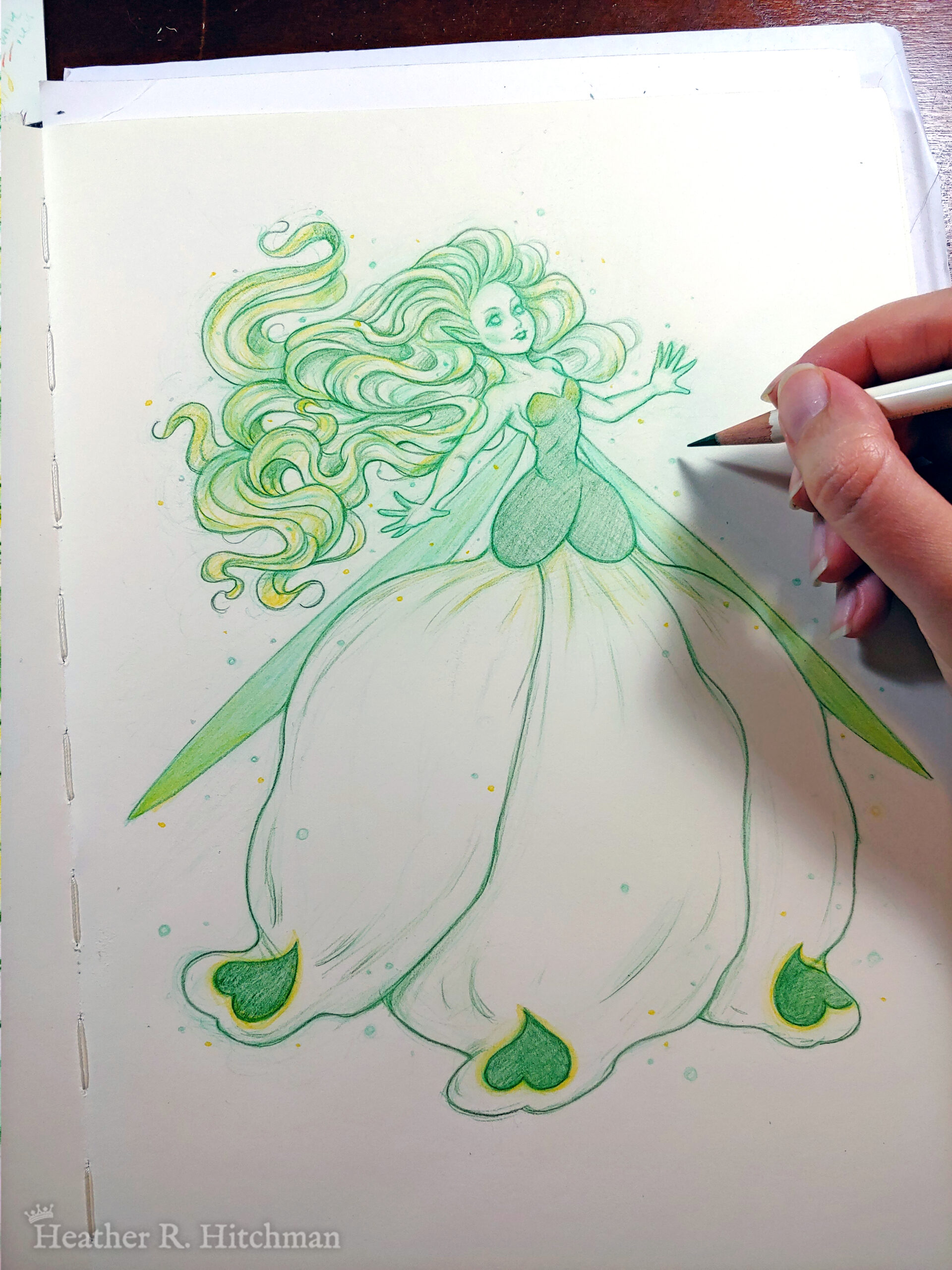 Photograph of a colored pencil full body drawing of a seelie snowdrop fae. The fae faces ¾ to the right, her arms outstretched to each side and her greenish white hair billowing behind her in a heap of floating curls. The fae wears a white and green ballgown shaped like a closed upside down snowdrop blossom. She has long narrow green wings that are angled downward behind her. The fae has a round childish face, with tiny black lips and large green eyes. She looks upward and to the right with a hopeful expression on her face. The hand of the artist is to the right holding a green pencil over the image.