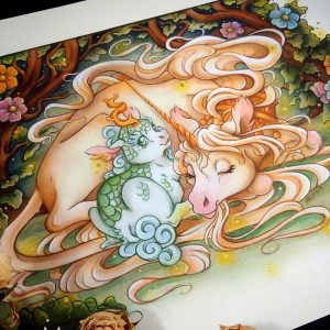 Photograph of a watercolor, gouache, and colored pencil painting print of a unicorn mother snuggling a green scaled baby kirin. The pair are nestled inside a thicket of green leaves and tree branches that arch overhead. Large orange, pink, and blue flowers emerge among the leaves. Gentle orbs of golden light float around the scene, and a soft glow of light radiates from the unicorn’s body. The unicorn is resting on the ground with her body facing to the right and her head curling back to meet the kirin nestling her back leg. She has light golden fur and appears to be sleeping with a knowing smile on her face. She has a long golden horn, a pink nose, and long tendrils of hair from her mane and fur floating around them. The unicorn also wears a golden crown around her neck. The baby kirin’s back is to the viewer as he sits right up against the unicorn. The baby kirin is looking back over his shoulder at the viewer with a joyful expression on his face. He has white fur, green scales across his back, and a soft blue cloud like mane and tail fur. The scene is depicted in a classic spot illustration style, with the outside edges of the background creating a border around the subject. The scene appears to float on a sepia backdrop. The painting rests against a black backdrop, with dried roses scattered over the painting to the bottom left.
