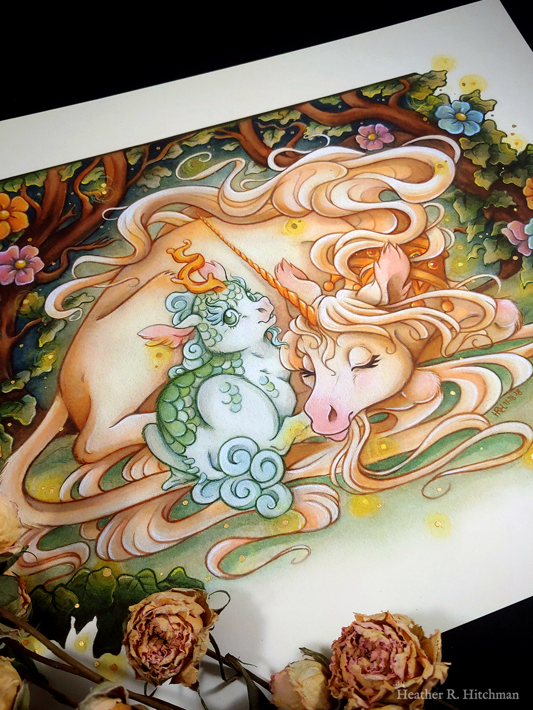 Photograph of a watercolor, gouache, and colored pencil painting print of a unicorn mother snuggling a green scaled baby kirin. The pair are nestled inside a thicket of green leaves and tree branches that arch overhead. Large orange, pink, and blue flowers emerge among the leaves. Gentle orbs of golden light float around the scene, and a soft glow of light radiates from the unicorn’s body. The unicorn is resting on the ground with her body facing to the right and her head curling back to meet the kirin nestling her back leg. She has light golden fur and appears to be sleeping with a knowing smile on her face. She has a long golden horn, a pink nose, and long tendrils of hair from her mane and fur floating around them. The unicorn also wears a golden crown around her neck. The baby kirin’s back is to the viewer as he sits right up against the unicorn. The baby kirin is looking back over his shoulder at the viewer with a joyful expression on his face. He has white fur, green scales across his back, and a soft blue cloud like mane and tail fur. The scene is depicted in a classic spot illustration style, with the outside edges of the background creating a border around the subject. The scene appears to float on a sepia backdrop. The painting rests against a black backdrop, with dried roses scattered over the painting to the bottom left.