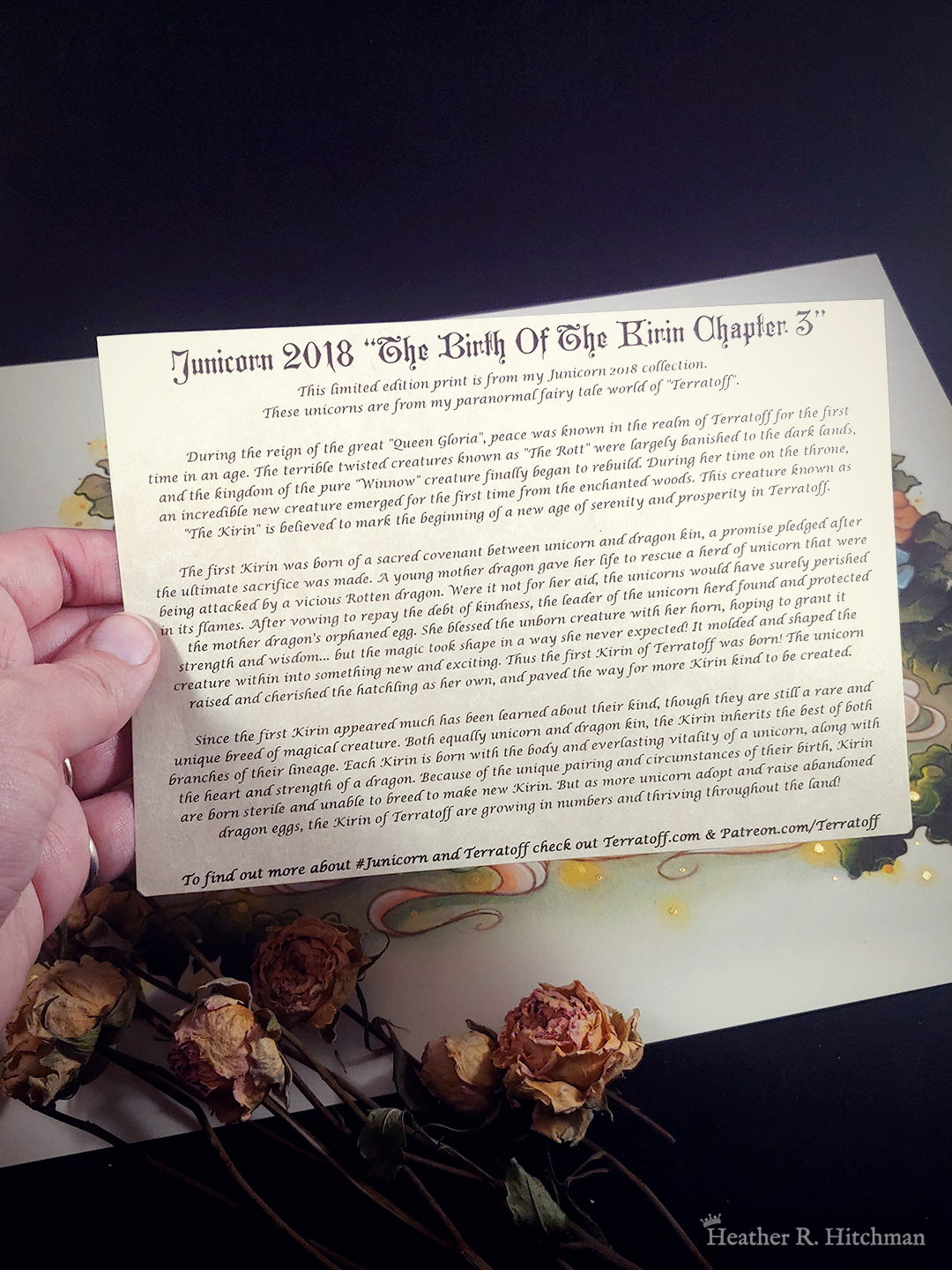 Photograph of the hand of the artist holding a “story scroll” over her painting “The Birth of the Terratoff Kirin.” The story scroll in a 5x7 inch parchment paper with a story printed on it in black ink in a whimsical font.