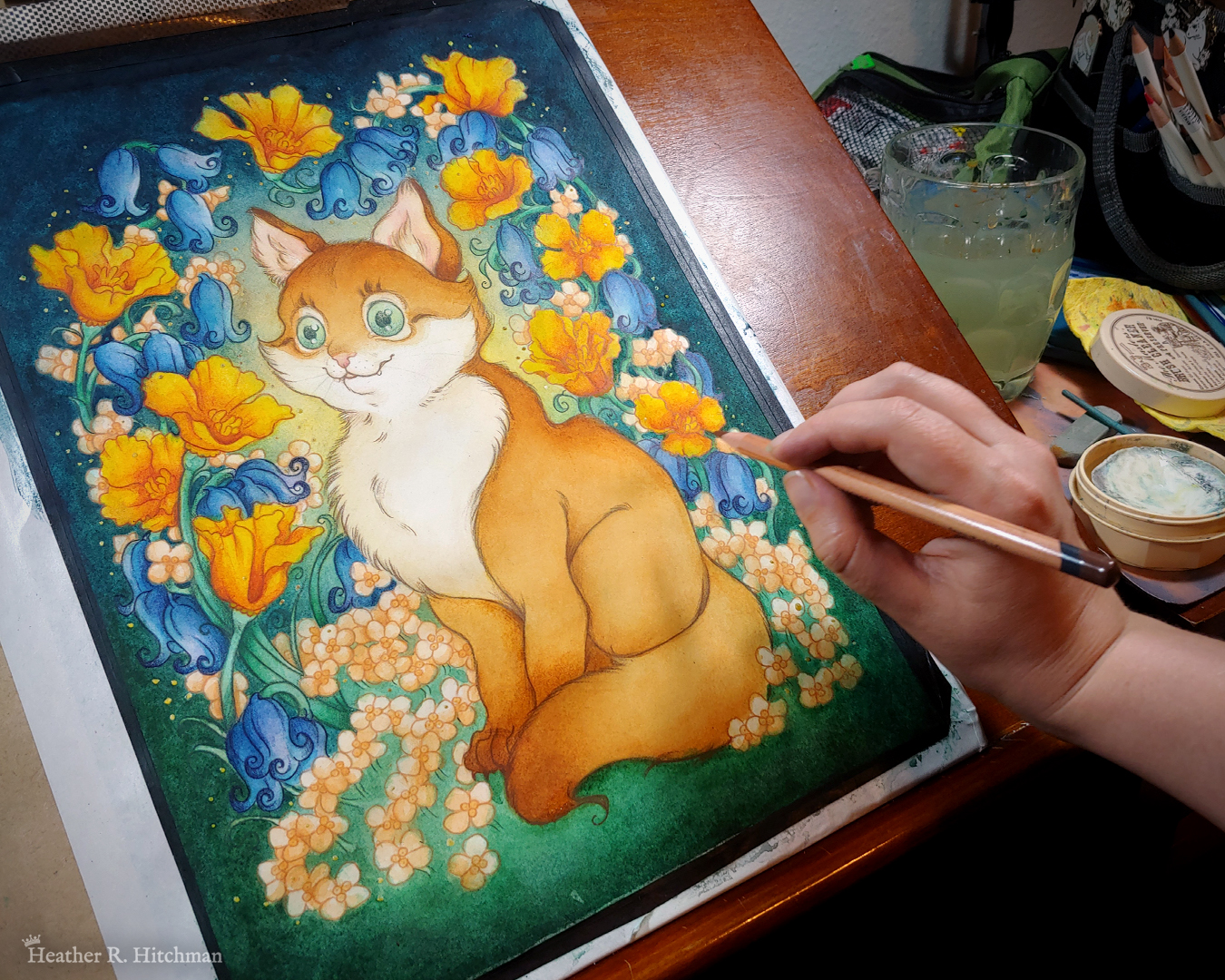 Photograph of a watercolor, gouache, and colored pencil portrait painting of an orange cat sitting in a garden. The painting is currently being painted by the artist, who’s hand is on the middle right edge of the photograph holding a colored pencil near right edge of the painting. The photograph shows the painting sitting on a wooden art desk, with art supplies sitting to the right behind the artist’s hand. The cat sits upright at a ¾ angle on the grass, facing to the left with a wide eyed happy expression on his face. His tail is coiled around in front of his feet. He has bright green eyes, white fur around his eyes and on his chest, and rich golden orange fur on the rest of his body. An archway of bluebells, yellow California poppies, and small white flowers surround the cat in an enchanted garden scene. The background is dark teal green with a sunburst of yellow light coming from behind the cat. So far the watercolor washes are loose and bright, with line art being added by the artist in colored pencil.
