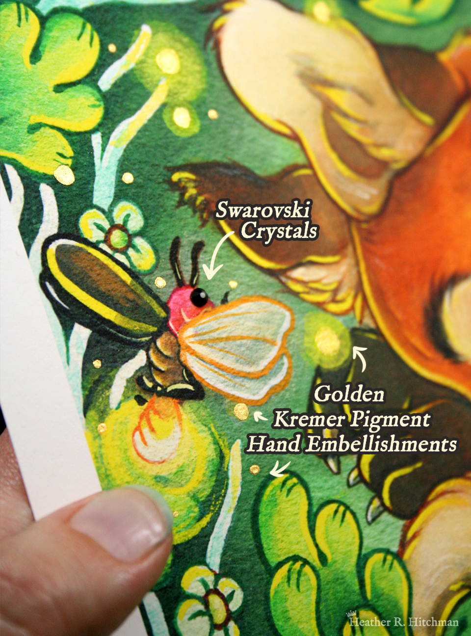 Close up photograph of a watercolor, gouache, ink, and colored pencil painting print of a sleeping fox with many tails known as a “kitsune”. The photograph focuses on the top left corner of the artwork titled “The Dreaming Kitsune” by Heather R. Hitchman. The hand of the artist is holding the print to the left besides a glowing firefly with a black swarovski crystal for its eye, and gold pigment hand painted on the glowing end of the insect. There is white text besides the firefly that reads “ Swarovski Crystals… Golden Kremer Pigment Hand Embellishments.”