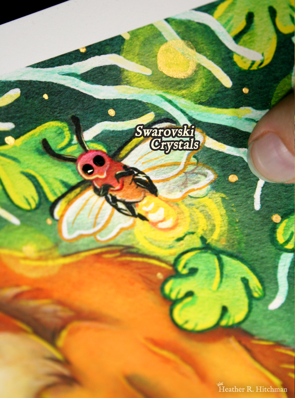 Close up photograph of a watercolor, gouache, ink, and colored pencil painting print of a sleeping fox with many tails known as a “kitsune”. The photograph focuses on the top right corner of the artwork titled “The Dreaming Kitsune” by Heather R. Hitchman. The hand of the artist is holding the print to the right besides a glowing firefly with black swarovski crystals for eyes, and gold pigment hand painted on the glowing end of the insect. There is white text besides the firefly that reads “ Swarovski Crystals”