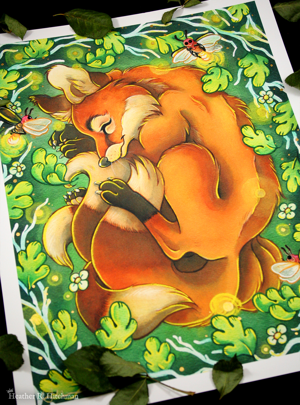 Photograph of a watercolor, gouache, ink, and colored pencil painting print of a sleeping fox with many tails known as a “kitsune”. The kitsune has golden orange fur, with white fur around his muzzle, eyes, and on the tips of his tails. His ears and paws have darker brown fur, and he appears to have 4 tails that are coiling around his feet. The kitsune is laying on his side with his front paws under his head like a pillow. He is coiling himself up in a cozy ball facing in profile to the left. The kitsune is sleeping with a tranquil expression on his face. He is laying on a green grass background surrounded by white branches and falling green leaves. Orbs of yellow light dance around the sleeping creature. 3 large fireflies are hovering in the scene, one in front of his face reaching for his ear, one by his shoulder, and the third by the base of his tails. Their rear ends are glowing with bright warm light. The print has a white border around it, and is surrounded by dried tree leaves.
