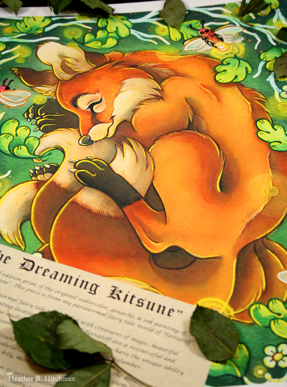 Photograph of a watercolor, gouache, ink, and colored pencil painting print of a sleeping fox with many tails known as a “kitsune”. The kitsune has golden orange fur, with white fur around his muzzle, eyes, and on the tips of his tails. His ears and paws have darker brown fur, and he appears to have 4 tails that are coiling around his feet. The kitsune is laying on his side with his front paws under his head like a pillow. He is coiling himself up in a cozy ball facing in profile to the left. The kitsune is sleeping with a tranquil expression on his face. He is laying on a green grass background surrounded by white branches and falling green leaves. Orbs of yellow light dance around the sleeping creature. 3 large fireflies are hovering in the scene, one in front of his face reaching for his ear, one by his shoulder, and the third by the base of his tails. Their rear ends are glowing with bright warm light. The print has a white border around it, and is surrounded by dried tree leaves. A scroll of parchment paper with a story printed on it in black in rests on top of the bottom left corner of the print.