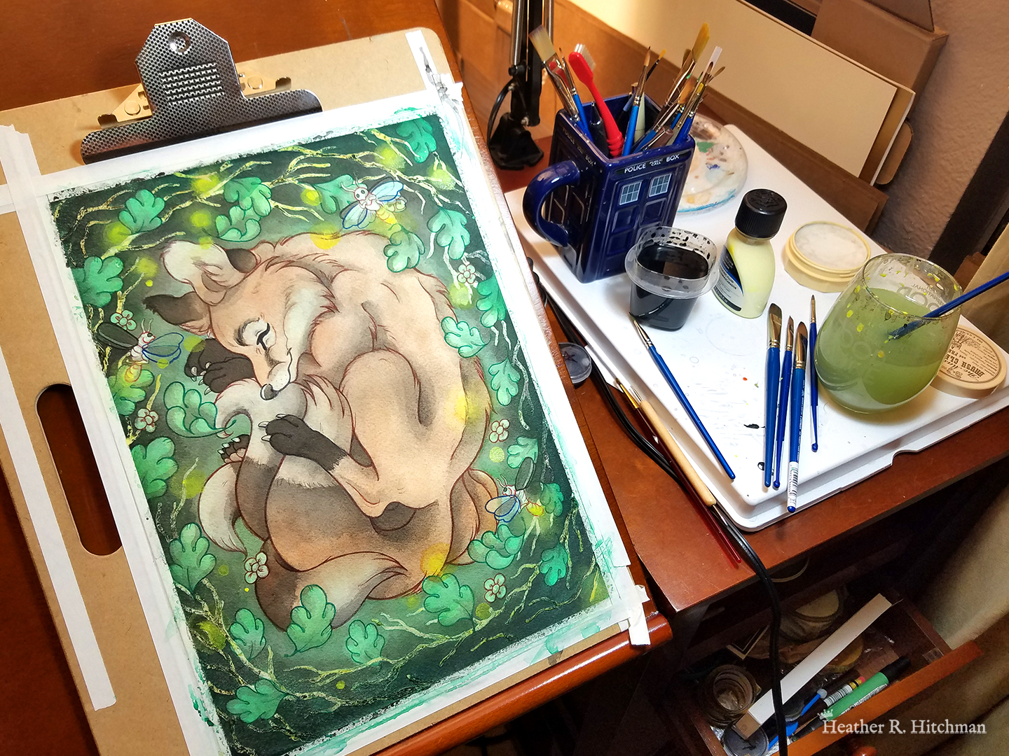 Photograph of a watercolor, gouache, and colored pencil painting of a sleeping fox with many tails known as a “kitsune”. The photograph is of the painting in progress, leaning on an art desk besides a tabletop of art supplies such as paint brushes, a cup of water, and a cup of ink. The kitsune has a light wash of golden orange fur, with white fur around his muzzle, eyes, and on the tips of his tails. His ears and paws have darker gray fur, and he appears to have 4 tails that are coiling around his feet. The kitsune is laying on his side with his front paws under his head like a pillow. He is coiling himself up in a cozy ball facing in profile to the left. The kitsune is sleeping with a tranquil expression on his face. He is laying on a green grass background surrounded by white branches and falling green leaves. Orbs of yellow light dance around the sleeping creature. 3 large fireflies are hovering in the scene, one in front of his face reaching for his ear, one by his shoulder, and the third by the base of his tails. Their rear ends are glowing with bright warm light.