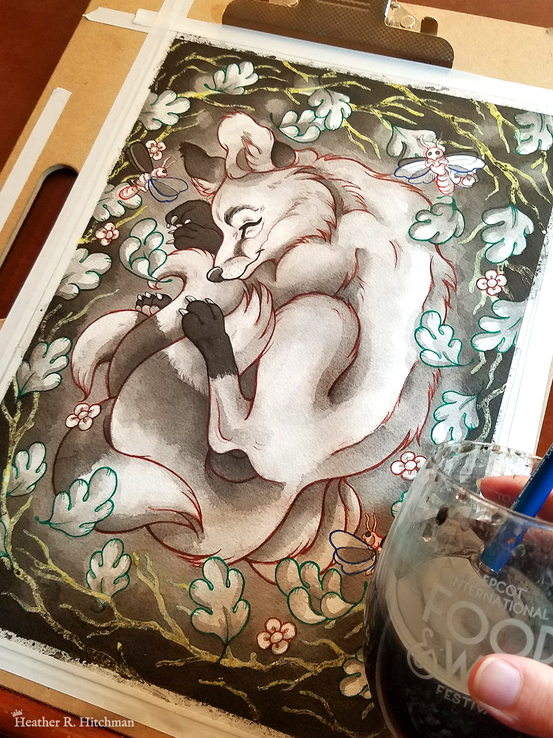 Photograph of a watercolor, gouache, and colored pencil painting of a sleeping fox with many tails known as a “kitsune”. The photograph is of the painting in progress, leaning on an art desk with the hand of the artist in the foreground holding a glass of inky water with a paintbrush inside. The artwork is currently line art and loose ink washes. The kitsune currently has white fur, with ears and paws of darker gray fur. He appears to have 4 tails that are coiling around his feet. The kitsune is laying on his side with his front paws under his head like a pillow. He is coiling himself up in a cozy ball facing in profile to the left. The kitsune is sleeping with a tranquil expression on his face. He is laying on a grass background surrounded by white branches and falling leaves. 3 large fireflies are hovering in the scene, one in front of his face reaching for his ear, one by his shoulder, and the third by the base of his tails.