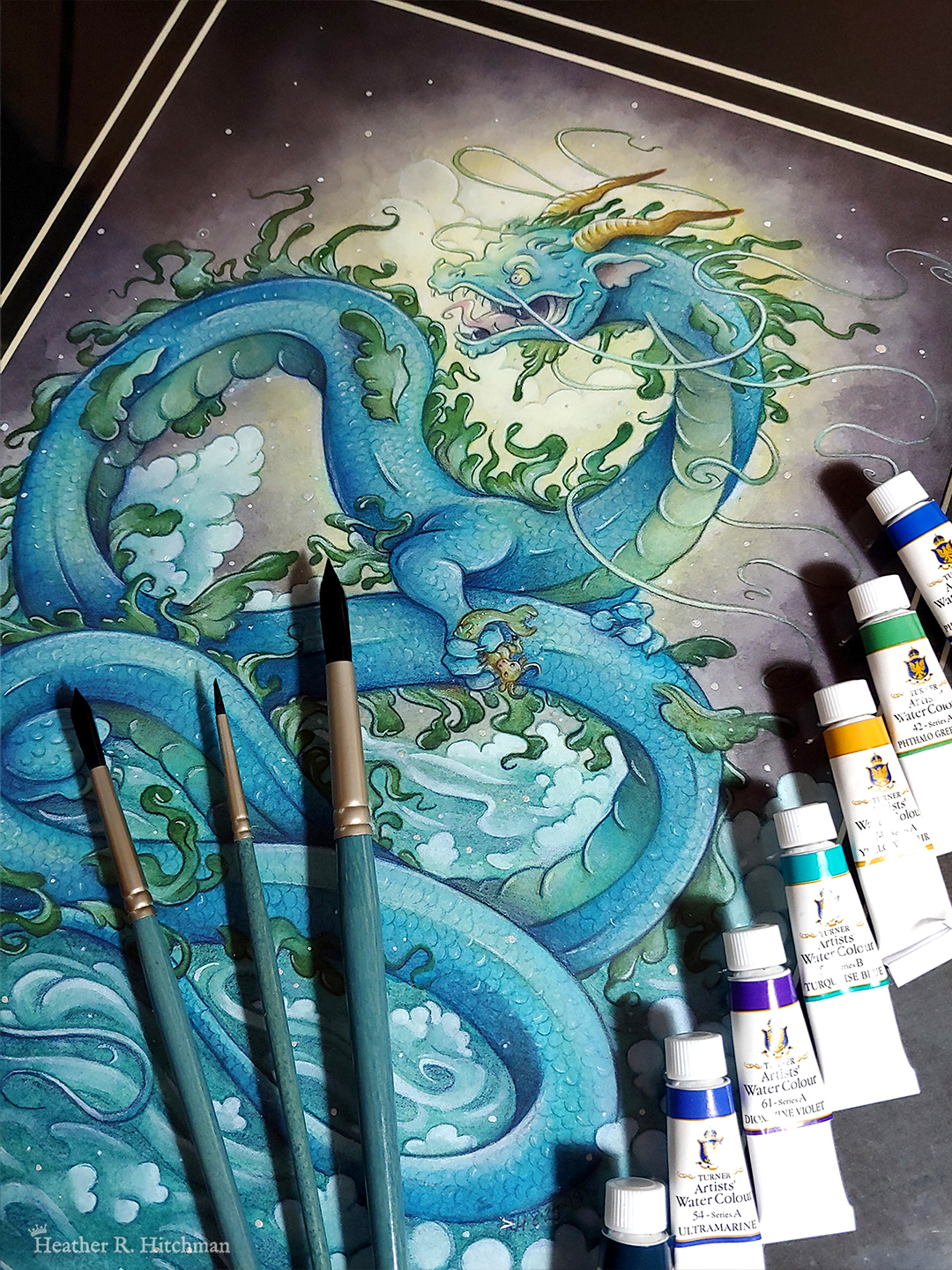 Photograph of a watercolor, gouache, and colored pencil painting of a gigantic water dragon sitting atop a sea of foaming waves out in the ocean. The sky behind the dragon is moody, almost completely covered with dark gray storm clouds. A burst of white and yellow sunlight breaks through the storm clouds behind the dragon’s head. The dragon has a long snakelike body covered in blue scales. Green frills of fins run down the length of the dragon’s back, and several more fins appear along its sides. The dragon has no wings, instead presenting like a classic eastern style dragon with long white whiskers, golden horns, front legs at the chest, and stumpy back legs towards the end of his long tail. The dragon’s body coils in a ribbon like shape, with its head upright and facing to the left. In it’s claws the dragon clutches a mer-bull that struggles to escape. The dragon’s face is menacing and mystical, with a hungry wicked smile showcasing his fangs and a giant pink tongue. The painting is matted in a double black mat, with a stack of watercolor paints resting on the right of the painting. 3 watercolor brushes are placed on top of the center bottom of the painting.